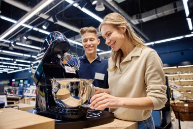 Meşgul bir perakende mağazasında, bilgili bir satış elemanı ilgili bir kadına yüksek teknoloji mutfak aletini göstererek, olumlu bir alışverişi teşvik etmek için benzersiz özelliklerini ve faydalarını vurgular.