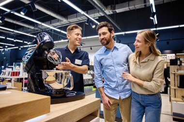 Meşgul bir perakende mağazasında, bilgili bir satış elemanı ilgili bir çifte yüksek teknoloji mutfak aletini göstererek, olumlu bir satın almayı teşvik etmek için eşsiz özelliklerini ve faydalarını vurgular.