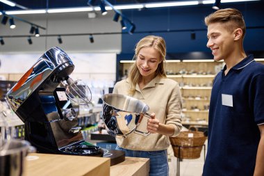 Meşgul bir perakende mağazasında, bilgili bir satış elemanı ilgili bir kadına yüksek teknoloji mutfak aletini göstererek, olumlu bir alışverişi teşvik etmek için benzersiz özelliklerini ve faydalarını vurgular.