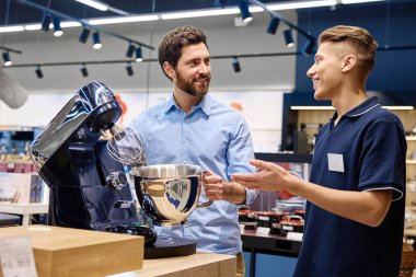 Bir müşteri, canlı bir perakende satış ortamı ile çevrili iken, bir prim standı mikserinin özelliklerini ve faydalarını keşfetmek için, bilgili bir satış danışmanıyla aktif bir şekilde sohbet eder.