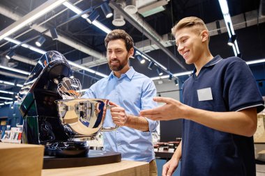 Bir müşteri, canlı bir perakende satış ortamı ile çevrili iken, bir prim standı mikserinin özelliklerini ve faydalarını keşfetmek için, bilgili bir satış danışmanıyla aktif bir şekilde sohbet eder.