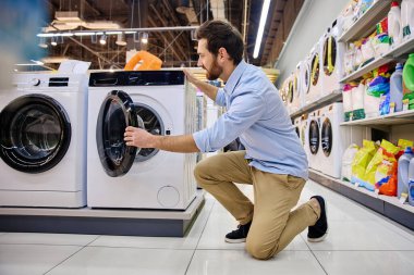 Müşteri, çeşitli çamaşır deterjanı seçenekleri ve sayısız temizlik ürünleriyle çevrili bir mağaza koridorunda bulunan şık ve modern bir çamaşır makinesini dikkatle inceliyor.