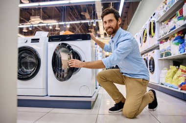 Müşteri, çeşitli çamaşır deterjanı seçenekleri ve sayısız temizlik ürünleriyle çevrili bir mağaza koridorunda bulunan şık ve modern bir çamaşır makinesini dikkatle inceliyor.