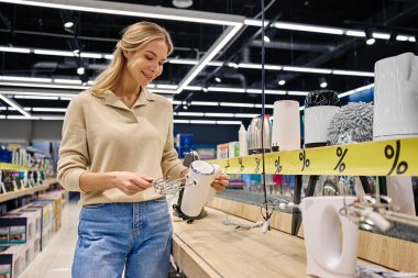 Neşeli genç bir kadın, şık bir mutfak aletini zevkle inceler. Parlak bir mağazada, hemen ilgisini çeken çeşitli düzenli ve indirimli ürünlerle dolu.