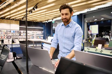 Moda bir elektronik butikte, genç bir adam çeşitli dizüstü bilgisayarları hevesle araştırıyor, modern teknolojiyi gözler önüne seriyor ve onların yenilikçi özellikleri ve işlevsellikleri için heyecanını dile getiriyor.