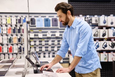Parlak mavi tişörtlü genç bir adam, hevesle yüksek teknoloji aksesuarlarına karşı ilgili alıcılara dizüstü bilgisayarlar ve tabletler gösteren hareketli bir teknoloji mağazasında çalışıyor.