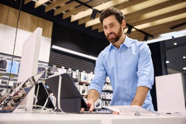 Parlak mavi tişörtlü genç bir adam, hevesle yüksek teknoloji aksesuarlarına karşı ilgili alıcılara dizüstü bilgisayarlar ve tabletler gösteren hareketli bir teknoloji mağazasında çalışıyor.