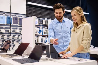 Çağdaş bir teknoloji mağazasında, arkadaş canlısı ve bilgili bir perakende temsilcisi müşteriye çeşitli ürünleri sergileyerek yardımcı olurken, aynı zamanda değerli rehberlik ve içgörü sunmaktadır.