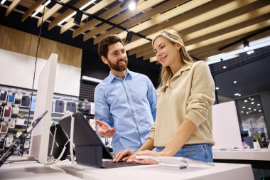 Çağdaş bir teknoloji mağazasında, arkadaş canlısı ve bilgili bir perakende temsilcisi müşteriye çeşitli ürünleri sergileyerek yardımcı olurken, aynı zamanda değerli rehberlik ve içgörü sunmaktadır.