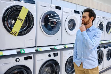 Bugünün modern perakende alışverişinin karmaşıklığını ve zorluklarını vurgulayan çeşitli seçenekleri ve fiyatları düşünceli bir şekilde tartan bir adam bir dizi çamaşır makinesinin önünde duruyor.