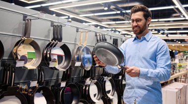 Bir adam, mutfak malzemesi satan bir mağazada sergilenen tavayı dikkatle inceliyor. Bu tavada, mutfak deneyimlerini geliştirmek için çeşitli mutfak gereçleri ve mutfak malzemesi seçenekleri sunuluyor.