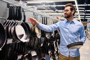 Bir adam, mutfak malzemesi satan bir mağazada sergilenen tavayı dikkatle inceliyor. Bu tavada, mutfak deneyimlerini geliştirmek için çeşitli mutfak gereçleri ve mutfak malzemesi seçenekleri sunuluyor.