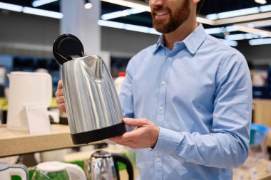 Çağdaş yemek meraklıları için tasarlanmış şık bir perakende mağazasında modern mutfak aletleriyle çevrili şık, parlak, paslanmaz çelik bir çaydanlığı dikkatle inceleyen bir adam.