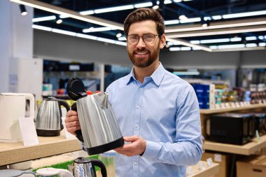 Çağdaş yemek meraklıları için tasarlanmış şık bir perakende mağazasında modern mutfak aletleriyle çevrili şık, parlak, paslanmaz çelik bir çaydanlığı dikkatle inceleyen bir adam.