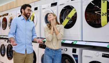 Yoğun bir mağazada, bir çift çamaşır makinelerini inceler, enerji verimliliği etiketlerini gözden geçirir ve çamaşır ihtiyaçlarına ve yaşam tarzlarına en uygun olanı bulmak için seçenekleri tartışır