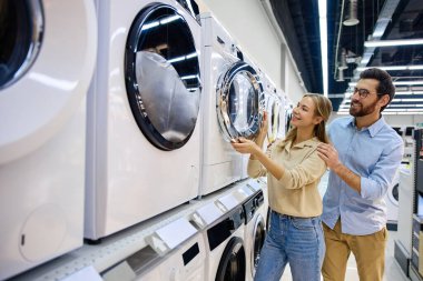 Genç bir çift, her bir model için farklı özellikleri, seçenekleri ve özellikleri tartışırken modern bir mağazada çeşitli çamaşır makinelerini hevesle keşfediyor.