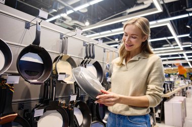 Gösterişli ve çağdaş bir perakende mağazasında, genç bir kadın çeşitli kızartma tavaları ve yemek takımlarına göz gezdiriyor, seçeneklerini dikkatlice değerlendiriyor ve seçeneklerini değerlendiriyor.
