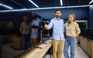 Çağdaş bir elektronik mağazasında, genç bir çift çeşitli televizyonlara göz gezdirirken piyasada çeşitli özellikleri ve seçenekleri tartışırken görülüyor.
