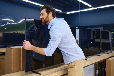 Bir adam, modern teknoloji yeniliklerine karşı coşkulu ve neşeli müşteri görüşlerini açıkça sergileyen bir elektronik mağazasında büyük bir düz ekran TV 'yi heyecanla kucaklarken görülebilir.