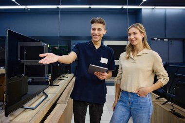 Çalışkan bir teknoloji mağazasında parlak ve neşeli bir etkileşimde, bilgili bir satıcı çeşitli televizyonlarda yardım arayan meraklı bir müşteriye uzman rehberliği sunar.