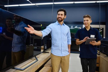 Çalışkan bir teknoloji mağazasında parlak ve neşeli bir etkileşimde, bilgili bir satıcı çeşitli televizyonlarda yardım arayan meraklı bir müşteriye uzman rehberliği sunar.