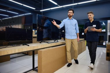 Çalışkan bir teknoloji mağazasında parlak ve neşeli bir etkileşimde, bilgili bir satıcı çeşitli televizyonlarda yardım arayan meraklı bir müşteriye uzman rehberliği sunar.