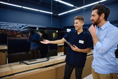 Çağdaş bir elektronik mağazasında, genç bir satış görevlisi, yüksek çözünürlüklü bir televizyonun etkileyici özelliklerini ve yeteneklerini ilgilenen bir müşteriye enerjik bir şekilde gösterir.