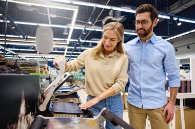 Modern ve iyi aydınlatılmış bir perakende mağazasında, neşeli bir çift, seçeneklerini keşfederken farklı mutfak aletleriyle coşkuyla etkileşime geçer, gülümseyerek ve birlikte eğlenerek.