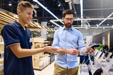 Çağdaş bir perakende satış ortamında, bir müşteri bir mağaza çalışanı ile alışverişe hazır çeşitli elektronik cihazları araştırırken sohbet eder.