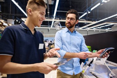 Çağdaş bir perakende satış ortamında, bir müşteri bir mağaza çalışanı ile alışverişe hazır çeşitli elektronik cihazları araştırırken sohbet eder.