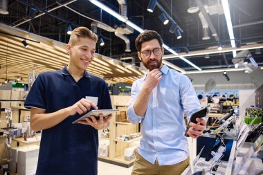 Çağdaş bir perakende satış ortamında, bir müşteri bir mağaza çalışanı ile alışverişe hazır çeşitli elektronik cihazları araştırırken sohbet eder.