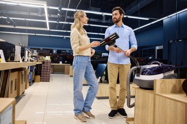 Genç bir çift, en son ev teknolojisini sergilemek üzere tasarlanmış modern bir perakende mağazasında gezinirken çeşitli elektrikli süpürgeleri ve diğer ev aletlerini aktif olarak değerlendiriyor.