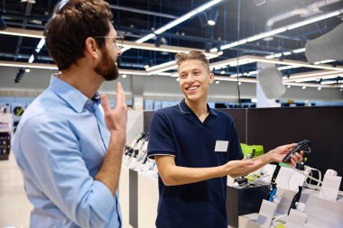 Çağdaş bir perakende satış ortamında, bir müşteri bir mağaza çalışanı ile alışverişe hazır çeşitli elektronik cihazları araştırırken sohbet eder.