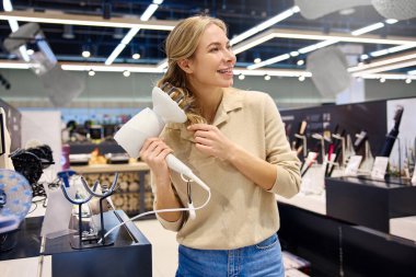 Neşeli bir kadın modern ve şık bir saç kurutma makinesini gelişmiş bir perakende ortamında test ediyor ve şu anda mevcut olan saç bakım teknolojisini gururla sergiliyor.