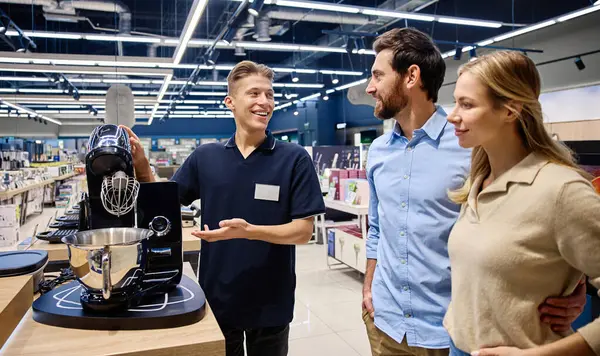 Meşgul bir perakende mağazasında, bilgili bir satış elemanı ilgili bir çifte yüksek teknoloji mutfak aletini göstererek, olumlu bir satın almayı teşvik etmek için eşsiz özelliklerini ve faydalarını vurgular.