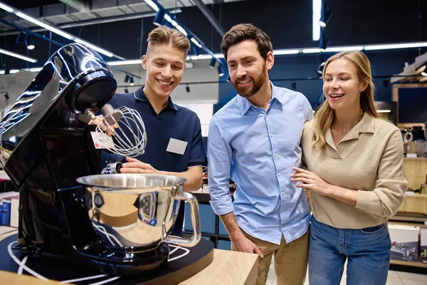 Meşgul bir perakende mağazasında, bilgili bir satış elemanı ilgili bir çifte yüksek teknoloji mutfak aletini göstererek, olumlu bir satın almayı teşvik etmek için eşsiz özelliklerini ve faydalarını vurgular.