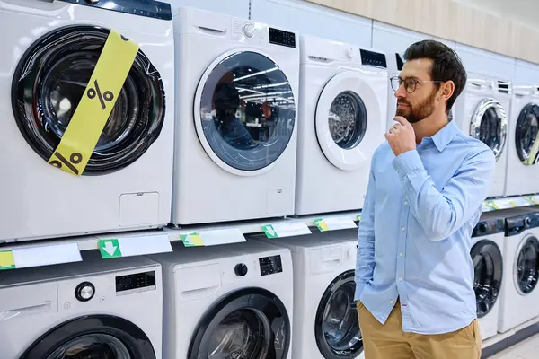 Bugünün modern perakende alışverişinin karmaşıklığını ve zorluklarını vurgulayan çeşitli seçenekleri ve fiyatları düşünceli bir şekilde tartan bir adam bir dizi çamaşır makinesinin önünde duruyor.