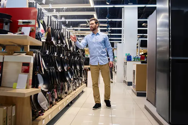 Gündelik kıyafet giymiş bir adam, çeşitli mutfak gereçleri ve mutfak gereçleri seçenekleri sunan çağdaş bir perakende mağazasında çok çeşitli mutfak gereçlerini incelerken görülüyor.