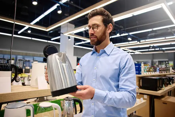 Çağdaş yemek meraklıları için tasarlanmış şık bir perakende mağazasında modern mutfak aletleriyle çevrili şık, parlak, paslanmaz çelik bir çaydanlığı dikkatle inceleyen bir adam.