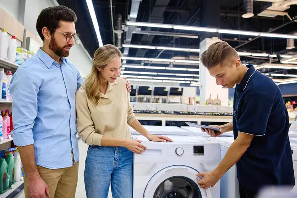 Bir çift, çağdaş bir elektronik mağazasında çeşitli çamaşır makinelerini araştırırken ve incelerken bilgili bir satıcıyla iletişim ve etkileşim halindedir.