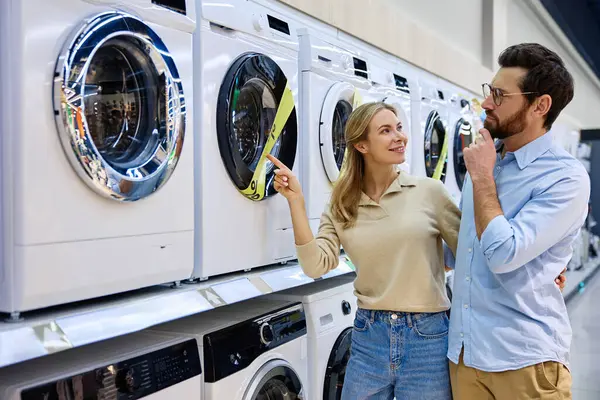 Genç bir çift, her bir model için farklı özellikleri, seçenekleri ve özellikleri tartışırken modern bir mağazada çeşitli çamaşır makinelerini hevesle keşfediyor.