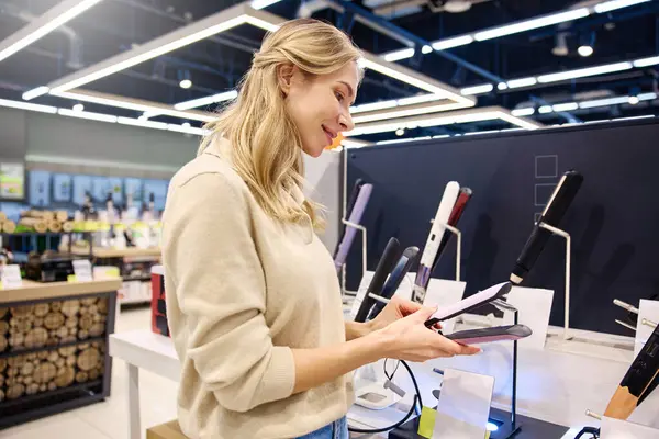 Genç bir kadın, modern ve şık bir perakende mağazasından uygun bir saç düzleştirici seçerken neşeli bir şekilde gülümsüyor. Etrafı canlı stil araçları ve modaya uygun dekorlarla çevrili.