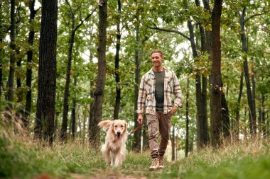 Bir adam, sevgili Golden Retriever 'ı ile birlikte canlı yeşillik ve doğanın güzelliği ile çevrili sakin bir orman patikasında huzurlu bir yürüyüşün keyfini çıkarmaktan büyük zevk alır.