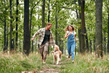 Neşeli ve mutlu bir çift coşkuyla neşeli, canlı, yemyeşil bir ormanda sevgili köpekleri eşliğinde koşar. Doğada değerli bir mutluluk ve bağ anını yakalar.
