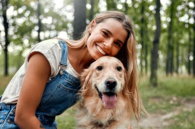 Neşeli genç bir kadın arkadaşlığın ve saf neşenin güzelliğinin altını çizerek, huzurlu ve huzurlu bir orman ortamında sevgili Golden Retriever 'ıyla eğleniyor.