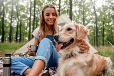 Neşeli bir kız, sakin ve sakin bir orman ortamında kamp yaparken Golden Retriever 'ıyla keyifli bir an yaşıyor ki bu da açık hava ve pettemalı içerik için ideal.