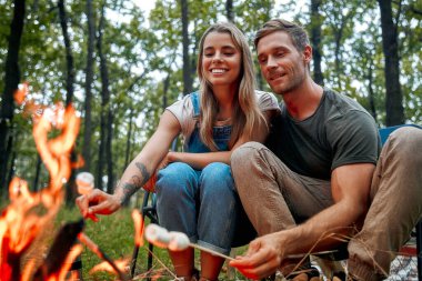 Neşeli bir çift, yıldızlı gece gökyüzünün güzelliğini kucaklarken birlikte geçirdikleri güzel zamanların tadını çıkararak, huzurlu ormandaki sıcak bir kamp ateşinde leziz marşmelovları kızartıyorlar.