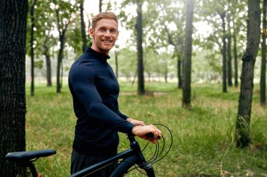 Canlı giysiler içinde neşeli bir bisikletçi lüks bir ormanla çevrili parlak bisikletinin yanında gülümsüyor. Macera dolu aktif bir yaşam tarzını ve doğa güzelliğine olan sevgisini simgeliyor.
