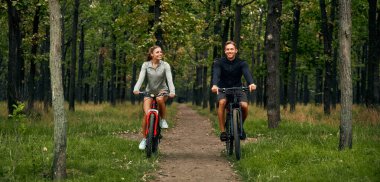 Neşeli bir çift, neşeli bir şekilde bisikletlerini sürüyorlar. Çevreleri canlı, yemyeşil bir orman patikasıyla çevrili. Açık hava ve macera temaları için mükemmel bir sahne.