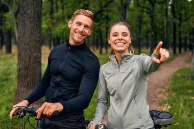 In the midst of their outdoor adventure, two cheerful and smiling cyclists take a delightful pause to savor the stunningly beautiful surroundings that nature has to offer during their journey